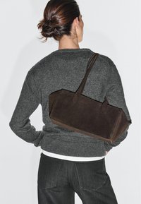 Frau mit dunklen Haaren zu einem Dutt gebunden, trägt einen grauen Pullover und dunkle Jeans, und hat eine große dunkelbraune Wildleder-Umhängetasche auf dem Rücken.