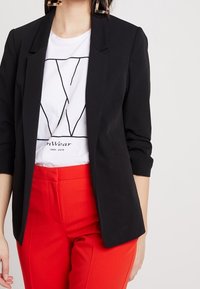 Czarna marynarka z klapami noszona na białym T-shircie z czarnym geometrycznym wzorem, w połączeniu z jaskrawoczerwonymi spodniami.