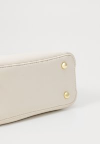 Esquina de un bolso de imitación de cuero beige que presenta dos acentos de tachuelas doradas, forma curva y costuras limpias a lo largo de los bordes. Se observa una textura suave.