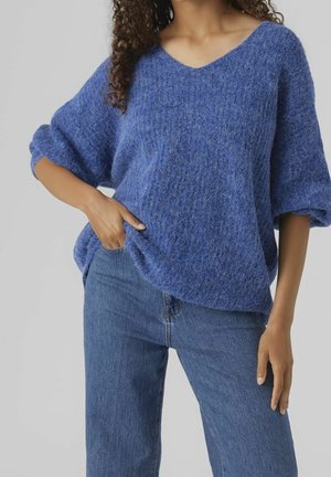 Vrouw draagt een blauw los textuurtrui met V-hals en opgerolde mouwen, gecombineerd met medium-wash blauwe spijkerbroek, hand in zak.