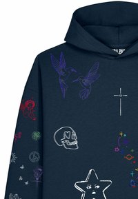 Sweat à capuche bleu marine avec des motifs brodés colorés incluant deux colibris, un crâne, une croix, une étoile avec un visage, et divers petits symboles sur la manche.