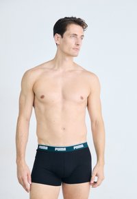 Schwarze Boxer-Shorts mit einem türkisfarbenen Bund, auf dem das "PUMA"-Logo zu sehen ist. Der Stoff wirkt glatt und dehnbar. Das Modell steht seitlich.