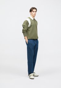 Sudadera verde oliva con cuello blanco, combinada con pantalones vaqueros azules. El conjunto presenta un ajuste relajado y zapatillas casuales.
