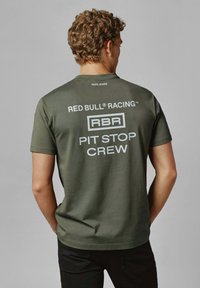 Olīvzaļš kokvilnas T-krekls ar apaļu kakla izgriezumu, aizmugurē ar baltu tekstu: "RED BULL® RACING," "RBR," un "PIT STOP CREW."