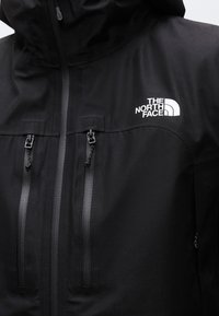 Veste d'extérieur noire avec capuche, deux poches poitrine verticales zippées et logo blanc "The North Face" sur le côté droit de la poitrine.