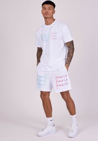 Conjunto de camiseta blanca y pantalones cortos con el texto colorido "Project Paris" en azul y rosa. Presenta un ajuste relajado, material de algodón y zapatillas deportivas.
