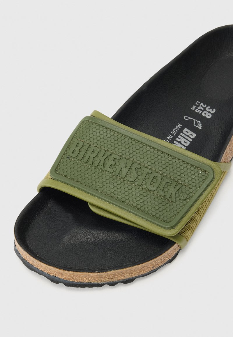 Sandale à semelle noire avec une lanière ajustable vert olive embossée avec "BIRKENSTOCK" et semelle intermédiaire en liège visible sur le côté.