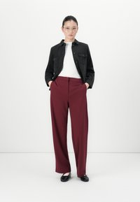 Woman wearing clear glasses, black jacket, white top, burgundy wide-leg trousers, and black ballet flats, standing with hands in pockets.