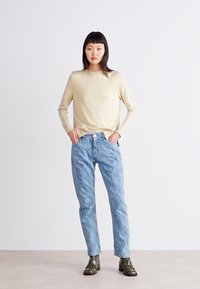 Top de manga longa bege, jeans azul claro com efeito marmoreado e botas de salto verdes. Modelo está em pé, com as mãos nos quadris, contra um fundo simples.