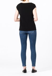 Kvinna står vänd bort, klädd i en svart kortärmad topp, blå tighta jeans och svarta slip-on skor mot en ren vit bakgrund.