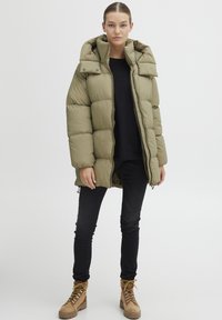 Olive green puffer jacket with a hood, featuring a zip front, large quilted sections, and snap buttons. Combined with black skinny jeans and tan boots.