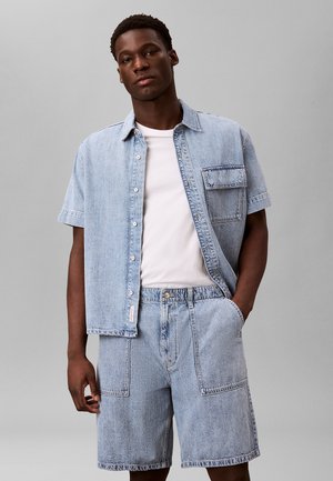 Mand iført lyseblå denim kortærmet skjorte over hvid t-shirt og matchende denimshorts med store forlommer, stående mod grå baggrund.