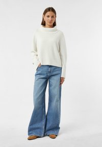 Weißer, grob gestrickter Pullover mit hohem, umgeschlagenem Kragen; kombiniert mit hellblauen, weit geschnittenen Jeans und braunen, strukturierten Stiefeletten.