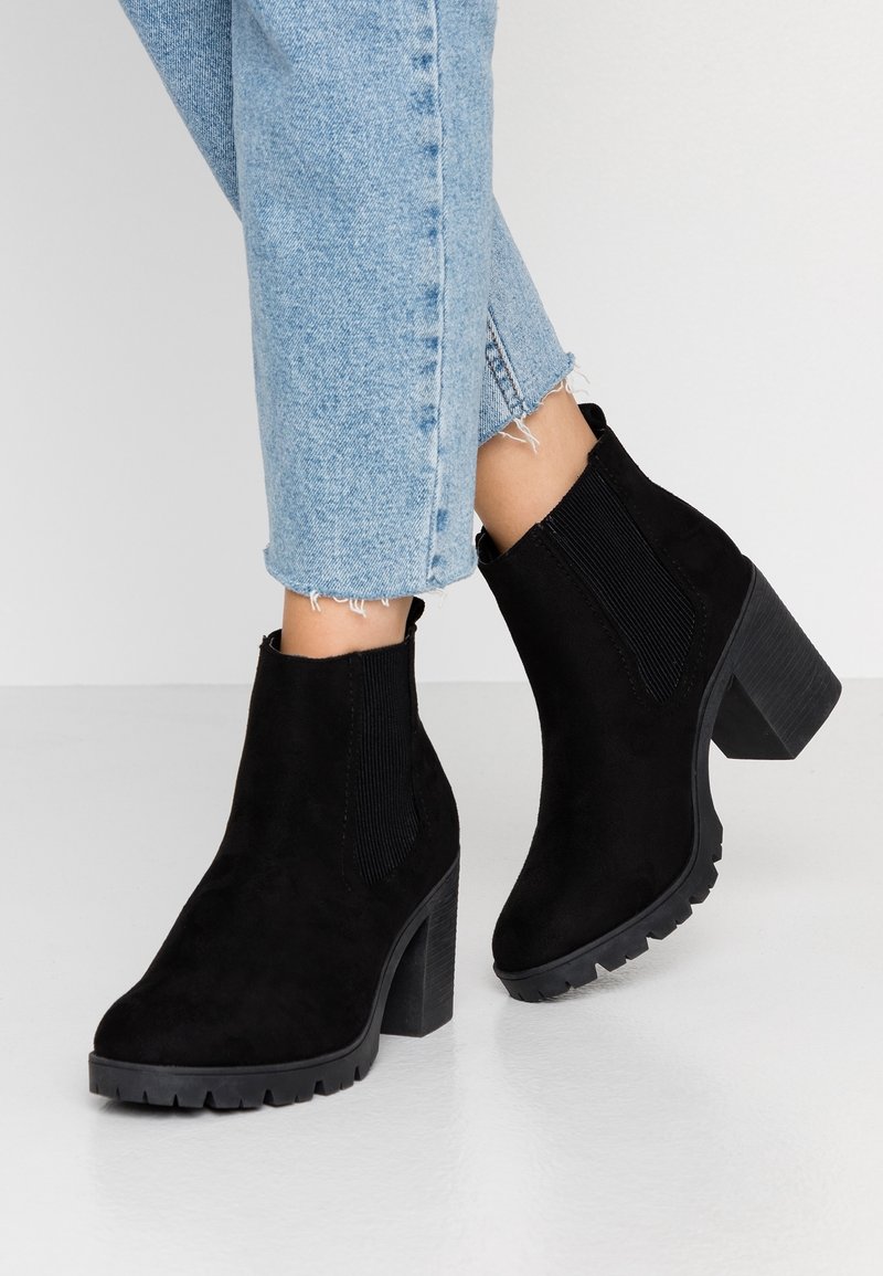 Bottines en daim noir avec talons épais portées avec un jean bleu clair court et effiloché, présentées sur fond blanc.