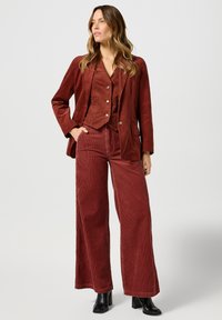 Costum din catifea în nuanță de roșu închis, având o jachetă ajustată cu nasturi și pantaloni cu picioare largi. Asortat cu cizme negre până la gleznă, punând în evidență nervurile verticale.