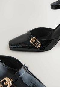 Black patent leather slingback heels with a square toe, featuring a gold-toned buckle detail on the strap and smooth textured surface.