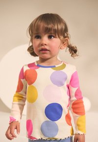 Dugi rukav rebrasti top s višebojnim kružnim uzorcima na krem pozadini, s kontrastnim plavim zaobljenim izrezom i naboranim rubom.