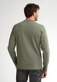 Cămașă lungă cu mâneci, de culoare verde măsliniu, dintr-un material moale, arătată din spate, combinată cu blugi închiși la culoare, cu detalii cusute pe buzunare.