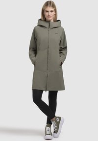 Giacca lunga verde oliva con cappuccio, cerniera frontale, tasche laterali e taglio dritto. Indossata sopra leggings neri e sneakers verdi.