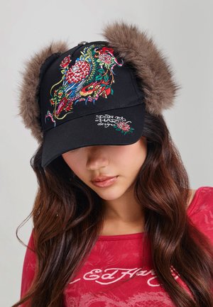 Mujer joven que lleva una gorra negra con bordado colorido de dragón y orejeras de piel marrón, y una camiseta roja de Ed Hardy.