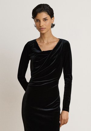 Femme aux cheveux courts et foncés portant une robe longue en velours noir à manches longues avec un décolleté drapé sur un fond uni.