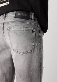 Jeans din denim gri cu un finisaj decolorat, având un patch din piele neagră cu logo-ul "BOSS" și un detaliu subtil de uzură aproape de buzunar.