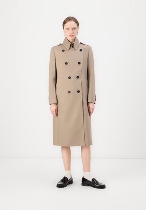 Trench coat beige doppiopetto con bottoni neri, tasche laterali e colletto. Il modello indossa mocassini neri e calzini chiari.