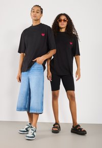 Black oversized t-shirts with heart and eye design, paired with wide blue jeans and black bike shorts. Footwear includes sneakers and sandals.