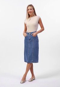 Γυναίκα φορώντας μια μπεζ χωρίς μανίκια μπλούζα, μπλε τζιν μίντι φούστα με τσέπες, και μεταλλικές χαμηλές γόβες, στέκεται με τα χέρια στις τσέπες.