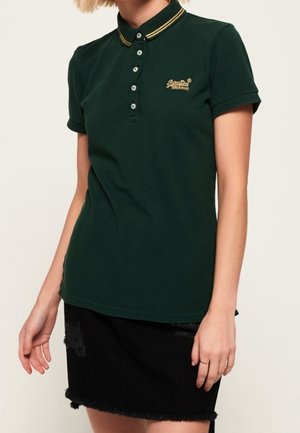 Femme portant un polo à manches courtes vert foncé avec un col rayé jaune et un logo brodé, assorti d'une jupe noire déchirée.
