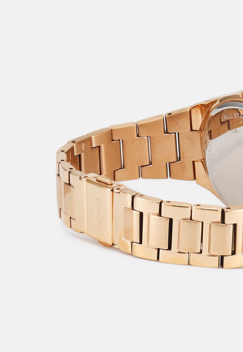 Montre en métal doré avec un bracelet à maillons structurés, présentant une finition polie. Le boîtier a une forme ronde avec un fond translucide.