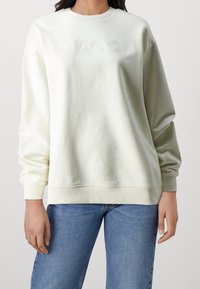 Persona che indossa una felpa off-white con delicata ricamo "HUGO" e jeans blu su uno sfondo chiaro e semplice.