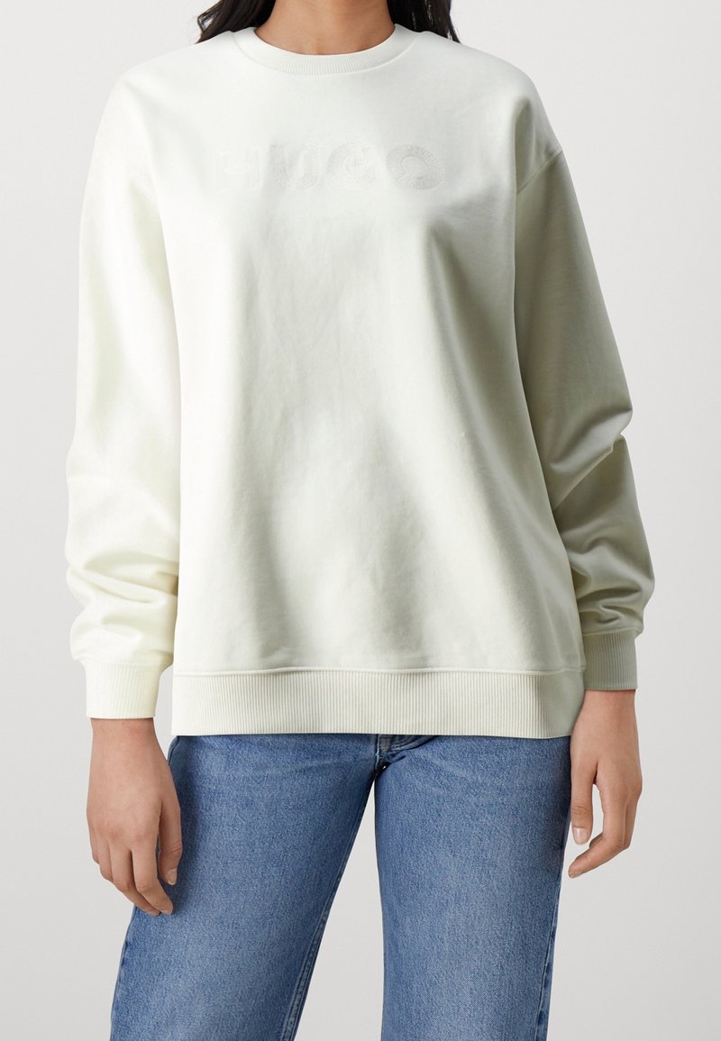 Persona che indossa una felpa off-white con delicata ricamo "HUGO" e jeans blu su uno sfondo chiaro e semplice.