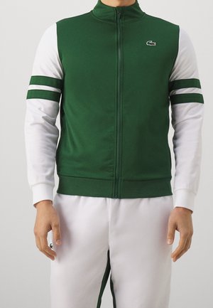 Homme portant un pantalon blanc et une veste zippée verte avec des manches blanches et deux rayures vertes sur chaque bras, debout devant un fond uni.