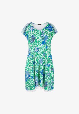 Femme portant une robe verte et blanche longueur genoux avec des motifs floraux et poissons bleus, associée à des baskets blanches et bleues.