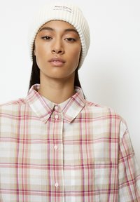 Plaid shirt with shades of pink, beige, and white. Featuring a button-down collar and a single breast pocket. A ribbed white beanie on the head.