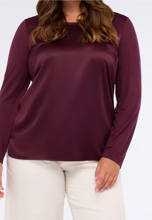 Vrouw draagt een lange mouw, donkerpaarse satijnen blouse en lichtbeige broek, staand tegen een effen lichte achtergrond.