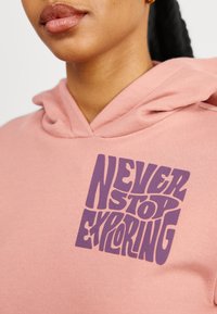 Gros plan d'une personne portant un sweat à capuche rose avec un texte violet sur la poitrine indiquant « Never Stop Exploring. »