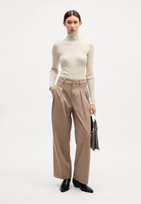 Bež pleten turtleneck pulover, kremne barve, v paru z visokim pasom in širokimi hlačami v kaki barvi. Model drži majhno srebrno torbico, črne čevlje.