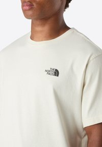 Muž oblečený v krémovém tričku s krátkým rukávem značky The North Face, na levém prsu s černým logem, na jednobarevném světlém pozadí.