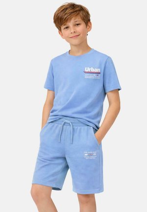 Jongen met kort bruin haar die een lichtblauw T-shirt en bijpassende korte broek draagt, handen in de zakken, licht glimlachend, tegen een effen achtergrond.