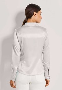 Frau mit dunkelhaarigem, nach hinten gebundenem Haar, die eine enganliegende, hellgraue, satinierte Bluse mit langen Ärmeln und weiße Hosen trägt, von der Kamera abgewandt.