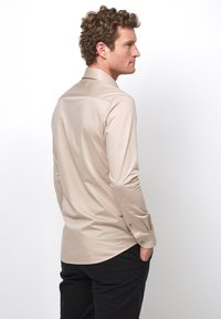 Helles beige Langarmhemd mit glatter Textur, ausgestattet mit Knopfmanschetten und einem leicht abgerundeten Saum. Auf dem Rücken eines Modells platziert.