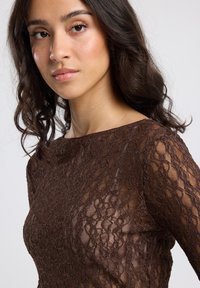 Vrouw met golvend donker haar, gekleed in een doorschijnende bruine kanten top, die rustig naar de camera kijkt tegen een effen lichte achtergrond.