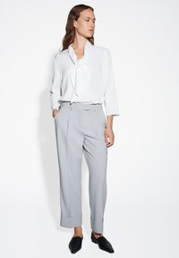 Weißbluse mit einer Schleifen-Detail, kombiniert mit hellgrauen, taillierten Hosen mit umgeschlagenen Säumen. Schwarze Slipper runden den Look ab.