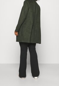 Lång, mörkgrön, faux mocka blazer med lös passform, kombinerad med svarta byxor och vita klackskor. Bakåtvyn visar en slät textur och design.