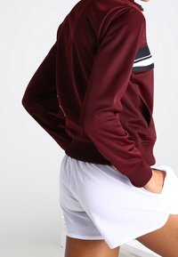 Bordeaux zipová bunda s bílými a černými vodorovnými pruhy na hrudi, spárovaná s bílými sportovními šortkami. Hladká, atletická tkanina.