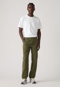 White short-sleeve t-shirt paired with olive green pants. Model stands with hands in pockets, wearing light gray and black sneakers.