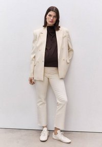 Beige skräddarsydd kavaj över en brun polotröja, med krämfärgade byxor och vita sneakers. Släta texturer; dubbelknäppt design med knappar.