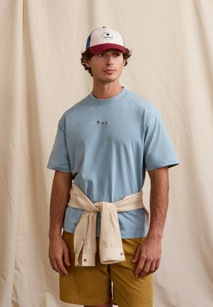 Jeune homme portant un t-shirt bleu, une veste beige nouée autour de la taille, un short moutarde et une casquette de baseball rouge, blanche et bleue, sur un fond beige.